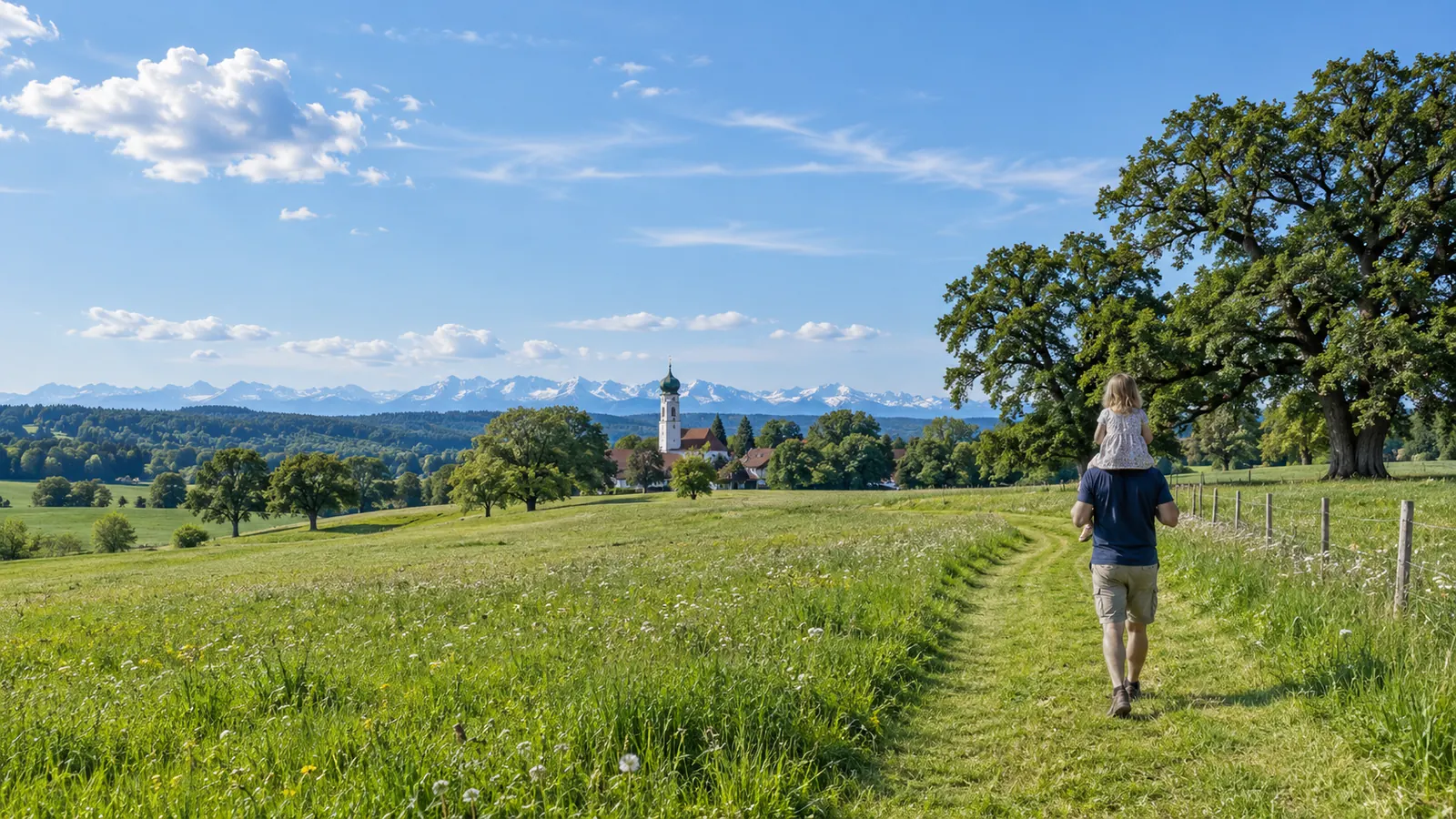 Ein Vater trägt seine Tochter auf den Schultern über eine sonnige Wiese, im Hintergrund Eichen, ein Kirchturm und die Alpen